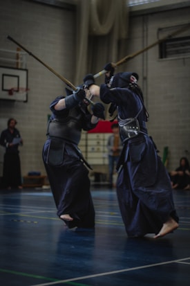 Two individuals wearing traditional kendo armor are engaged in a practice or match on an indoor court. They are holding long shinai swords, appearing to be in the midst of a sparring session. The setting is a sports hall with a basketball hoop visible in the background.