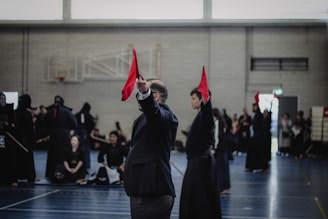 A martial arts event taking place in an indoor sports hall. Several individuals dressed in traditional martial arts attire participate, some wearing protective gear. A person prominently holds up a red flag, potentially indicating a referee or judge in action. The background includes spectators and sports equipment.