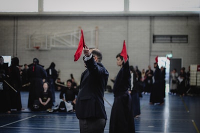 A martial arts event taking place in an indoor sports hall. Several individuals dressed in traditional martial arts attire participate, some wearing protective gear. A person prominently holds up a red flag, potentially indicating a referee or judge in action. The background includes spectators and sports equipment.
