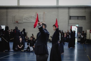 A martial arts event taking place in an indoor sports hall. Several individuals dressed in traditional martial arts attire participate, some wearing protective gear. A person prominently holds up a red flag, potentially indicating a referee or judge in action. The background includes spectators and sports equipment.