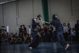 Two individuals wearing kendo armor are engaged in a sparring match on an indoor court. They hold bamboo practice swords and are surrounded by a group of onlookers, some seated and others standing, observing the action. The setting appears to be a gymnasium with brick walls and a high ceiling.