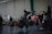 Two individuals in traditional kendo attire engage in a sparring match in an indoor setting, observed by a group of spectators sitting and standing nearby. The room has a large brick wall, and several people are capturing the event, possibly with cameras.