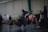 Two individuals in traditional kendo attire engage in a sparring match in an indoor setting, observed by a group of spectators sitting and standing nearby. The room has a large brick wall, and several people are capturing the event, possibly with cameras.