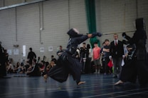 Two individuals in traditional kendo attire engage in a sparring match in an indoor setting, observed by a group of spectators sitting and standing nearby. The room has a large brick wall, and several people are capturing the event, possibly with cameras.