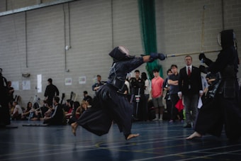 Two individuals in traditional kendo attire engage in a sparring match in an indoor setting, observed by a group of spectators sitting and standing nearby. The room has a large brick wall, and several people are capturing the event, possibly with cameras.