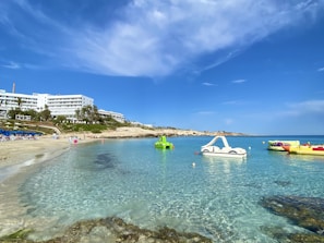 A serene beach scene with crystal-clear turquoise water and a sandy shore. Several white hotel buildings line the background, partially obscured by lush greenery and palm trees. Pedal boats and water inflatables are floating on the calm sea, under a bright blue sky with scattered clouds.