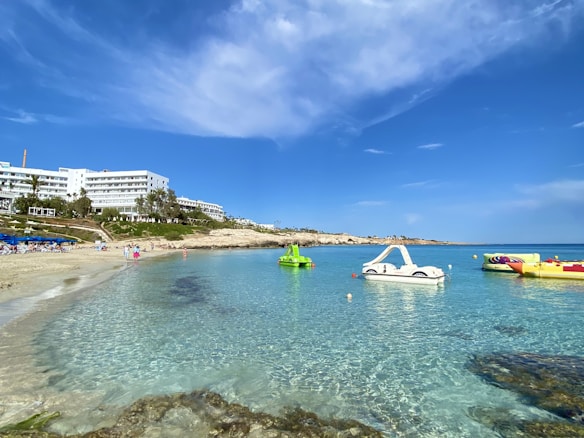 A serene beach scene with crystal-clear turquoise water and a sandy shore. Several white hotel buildings line the background, partially obscured by lush greenery and palm trees. Pedal boats and water inflatables are floating on the calm sea, under a bright blue sky with scattered clouds.