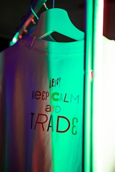 A T-shirt is hanging on a white hanger illuminated by colorful lights. The shirt features text in various fonts and colors, partially reading 'KEEP CALM AND TRADE.' The scene has a modern and vibrant feel due to the mix of green and red light reflections.