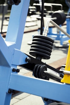 A close-up view of gym equipment focusing on a load of stacked weight plates on a metal rod. The equipment is primarily made from blue metal with black bolts and a yellow element in the corner. The background includes other gym machines and outdoor flooring.