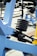 A close-up view of gym equipment focusing on a load of stacked weight plates on a metal rod. The equipment is primarily made from blue metal with black bolts and a yellow element in the corner. The background includes other gym machines and outdoor flooring.