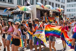 A vibrant parade scene with people of various ages walking along a city street. Many are holding rainbow flags and wearing colorful clothing. There is a decorated truck with umbrellas and a stuffed animal on top. The atmosphere is lively and festive.