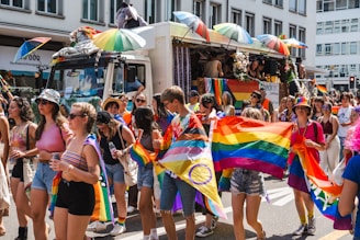 A vibrant parade scene with people of various ages walking along a city street. Many are holding rainbow flags and wearing colorful clothing. There is a decorated truck with umbrellas and a stuffed animal on top. The atmosphere is lively and festive.