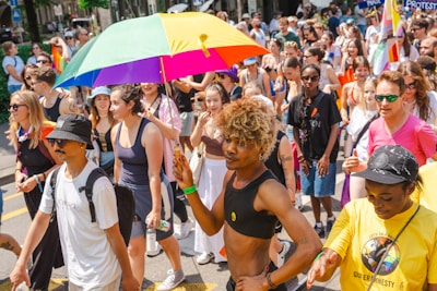 A large group of people participating in a street parade, with one individual prominently holding a colorful rainbow umbrella. The crowd is diverse, dressed in bright and casual attire, with some people wearing sunglasses and hats. Many appear to be celebrating and enjoying the event.
