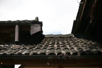 A close-up view of a weathered, tiled roof with dark, textured tiles. There is a pipe protruding from the structure, and dry leaves and debris are scattered across the tiles. The roof shows signs of aging and exposure to the elements, with some spots appearing mossy or worn.