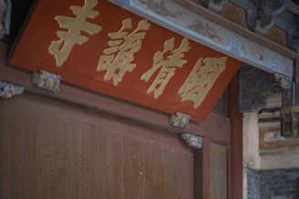 An ornate wooden structure features intricate carvings and a central red plaque with gold Chinese characters. The plaque is surrounded by decorative elements and traditional architectural details, conveying a sense of cultural heritage.