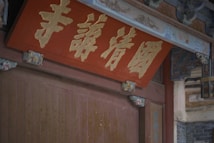 An ornate wooden structure features intricate carvings and a central red plaque with gold Chinese characters. The plaque is surrounded by decorative elements and traditional architectural details, conveying a sense of cultural heritage.