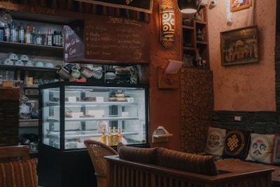 A cozy cafe interior with a refrigerated display case filled with desserts and beverages. The walls are adorned with eclectic decorations including framed art and shelves holding various bottles and cups. There is a small chalkboard listing the day's specials. Comfortable seating and warm lighting create an inviting atmosphere.