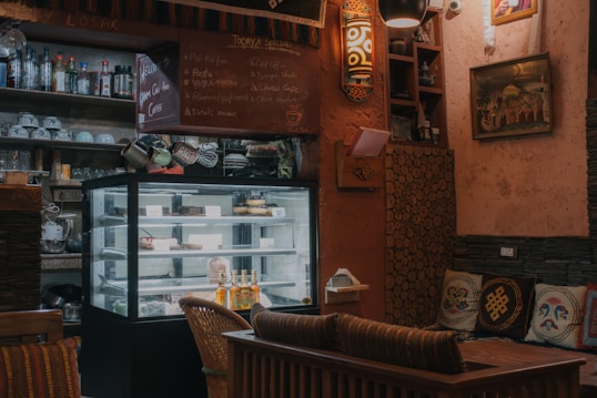 A cozy cafe interior with a refrigerated display case filled with desserts and beverages. The walls are adorned with eclectic decorations including framed art and shelves holding various bottles and cups. There is a small chalkboard listing the day's specials. Comfortable seating and warm lighting create an inviting atmosphere.
