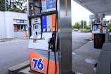 A gas station with multiple fuel pumps, each equipped with digital displays and labeled with different fuel options and prices. A convenience store is visible in the background, along with signage displaying promotions and prices. The parking area is spacious, surrounded by some greenery and parked vehicles.