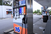 A gas station with multiple fuel pumps, each equipped with digital displays and labeled with different fuel options and prices. A convenience store is visible in the background, along with signage displaying promotions and prices. The parking area is spacious, surrounded by some greenery and parked vehicles.