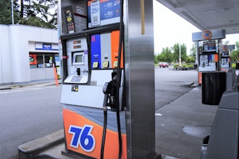 A gas station with multiple fuel pumps, each equipped with digital displays and labeled with different fuel options and prices. A convenience store is visible in the background, along with signage displaying promotions and prices. The parking area is spacious, surrounded by some greenery and parked vehicles.