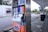 A gas station with multiple fuel pumps, each equipped with digital displays and labeled with different fuel options and prices. A convenience store is visible in the background, along with signage displaying promotions and prices. The parking area is spacious, surrounded by some greenery and parked vehicles.