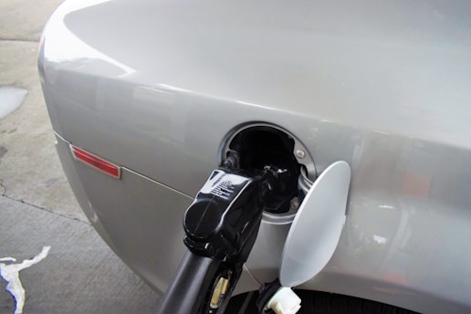 An automobile is being refueled at a gas station, with a visible fuel nozzle inserted into the vehicle's gas tank. The gas cap is open, and the car's exterior is silver.