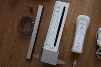 A set of gaming accessories placed on a wooden surface, including a gaming console standing vertically, accompanied by a controller, a sensor bar, and wires.