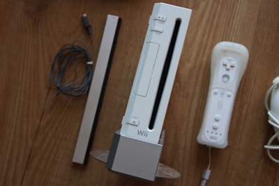 A set of gaming accessories placed on a wooden surface, including a gaming console standing vertically, accompanied by a controller, a sensor bar, and wires.