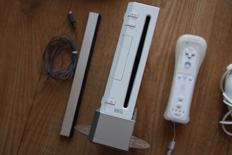 A set of gaming accessories placed on a wooden surface, including a gaming console standing vertically, accompanied by a controller, a sensor bar, and wires.