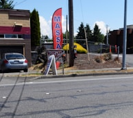 A gray car is parked in front of a one-story building with a horizontal maroon stripe across the top. A tall red feather flag with 'MASSAGE' in blue letters is next to a smaller A-frame sign also advertising a massage. The scene includes a paved driveway and a street with some visible road markings. In the background, there are trees, bushes, and power poles, all under a clear blue sky.