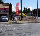 A gray car is parked in front of a one-story building with a horizontal maroon stripe across the top. A tall red feather flag with 'MASSAGE' in blue letters is next to a smaller A-frame sign also advertising a massage. The scene includes a paved driveway and a street with some visible road markings. In the background, there are trees, bushes, and power poles, all under a clear blue sky.