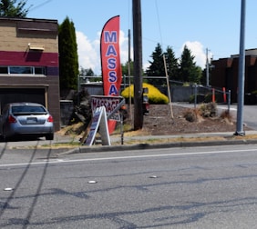 A gray car is parked in front of a one-story building with a horizontal maroon stripe across the top. A tall red feather flag with 'MASSAGE' in blue letters is next to a smaller A-frame sign also advertising a massage. The scene includes a paved driveway and a street with some visible road markings. In the background, there are trees, bushes, and power poles, all under a clear blue sky.