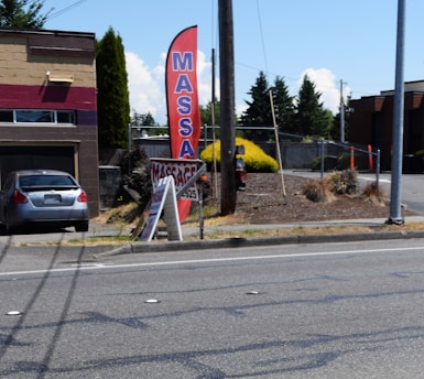 A gray car is parked in front of a one-story building with a horizontal maroon stripe across the top. A tall red feather flag with 'MASSAGE' in blue letters is next to a smaller A-frame sign also advertising a massage. The scene includes a paved driveway and a street with some visible road markings. In the background, there are trees, bushes, and power poles, all under a clear blue sky.