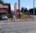 A gray car is parked in front of a one-story building with a horizontal maroon stripe across the top. A tall red feather flag with 'MASSAGE' in blue letters is next to a smaller A-frame sign also advertising a massage. The scene includes a paved driveway and a street with some visible road markings. In the background, there are trees, bushes, and power poles, all under a clear blue sky.