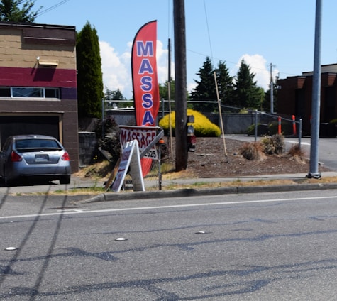 A gray car is parked in front of a one-story building with a horizontal maroon stripe across the top. A tall red feather flag with 'MASSAGE' in blue letters is next to a smaller A-frame sign also advertising a massage. The scene includes a paved driveway and a street with some visible road markings. In the background, there are trees, bushes, and power poles, all under a clear blue sky.