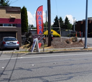A gray car is parked in front of a one-story building with a horizontal maroon stripe across the top. A tall red feather flag with 'MASSAGE' in blue letters is next to a smaller A-frame sign also advertising a massage. The scene includes a paved driveway and a street with some visible road markings. In the background, there are trees, bushes, and power poles, all under a clear blue sky.