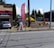 A gray car is parked in front of a one-story building with a horizontal maroon stripe across the top. A tall red feather flag with 'MASSAGE' in blue letters is next to a smaller A-frame sign also advertising a massage. The scene includes a paved driveway and a street with some visible road markings. In the background, there are trees, bushes, and power poles, all under a clear blue sky.
