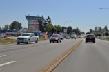 A busy road with several vehicles, including trucks and cars, driving in both directions. On the side of the road, there is a sign for Parkland Centre with several business names listed. Surrounding the road are trees and various commercial buildings.