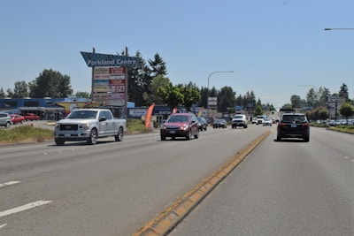 A busy road with several vehicles, including trucks and cars, driving in both directions. On the side of the road, there is a sign for Parkland Centre with several business names listed. Surrounding the road are trees and various commercial buildings.