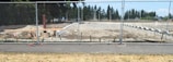 A construction site is surrounded by a chain-link fence, with a cleared area of dirt and some construction materials and equipment scattered around. In the background, there are trees and a partly cloudy sky.