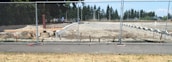A construction site is surrounded by a chain-link fence, with a cleared area of dirt and some construction materials and equipment scattered around. In the background, there are trees and a partly cloudy sky.