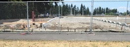 A construction site is surrounded by a chain-link fence, with a cleared area of dirt and some construction materials and equipment scattered around. In the background, there are trees and a partly cloudy sky.
