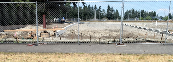 A construction site is surrounded by a chain-link fence, with a cleared area of dirt and some construction materials and equipment scattered around. In the background, there are trees and a partly cloudy sky.