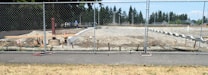 A construction site is surrounded by a chain-link fence, with a cleared area of dirt and some construction materials and equipment scattered around. In the background, there are trees and a partly cloudy sky.