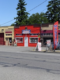 A small massage parlor with a bright red facade is located along a street. It features a prominent blue and white sign with the word 'MASSAGE' above the entrance. There is a large red vertical banner with the same word on the sidewalk. Several potted plants and decorations are placed near the door.
