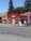 A small massage parlor with a bright red facade is located along a street. It features a prominent blue and white sign with the word 'MASSAGE' above the entrance. There is a large red vertical banner with the same word on the sidewalk. Several potted plants and decorations are placed near the door.