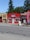 A small massage parlor with a bright red facade is located along a street. It features a prominent blue and white sign with the word 'MASSAGE' above the entrance. There is a large red vertical banner with the same word on the sidewalk. Several potted plants and decorations are placed near the door.