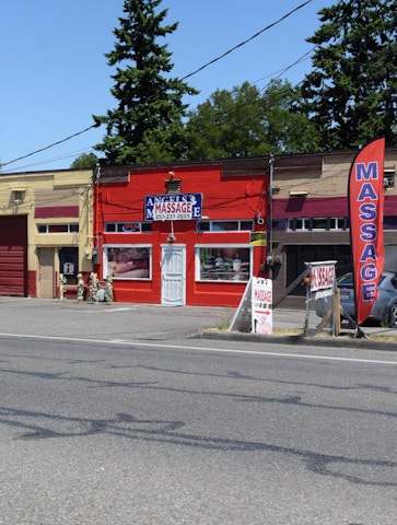 A small massage parlor with a bright red facade is located along a street. It features a prominent blue and white sign with the word 'MASSAGE' above the entrance. There is a large red vertical banner with the same word on the sidewalk. Several potted plants and decorations are placed near the door.