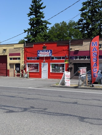 A small massage parlor with a bright red facade is located along a street. It features a prominent blue and white sign with the word 'MASSAGE' above the entrance. There is a large red vertical banner with the same word on the sidewalk. Several potted plants and decorations are placed near the door.